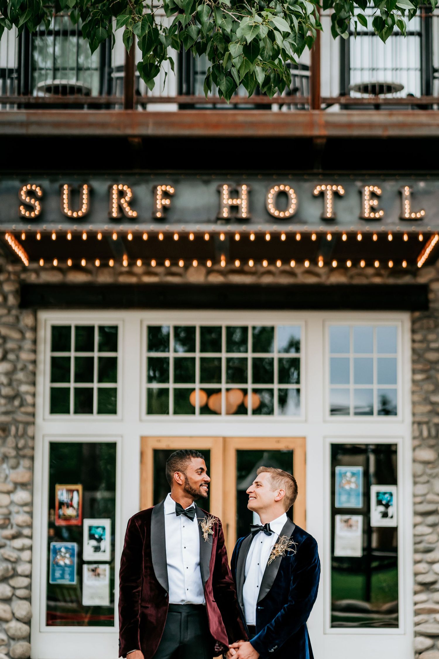 Couple poses in front of illuminated Surf Hotel sign on stone building facade.