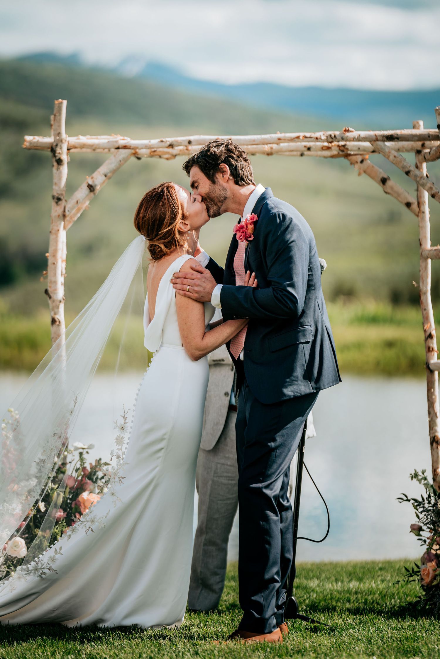 Wedding ceremony under rustic wooden arch with mountain lake backdrop.