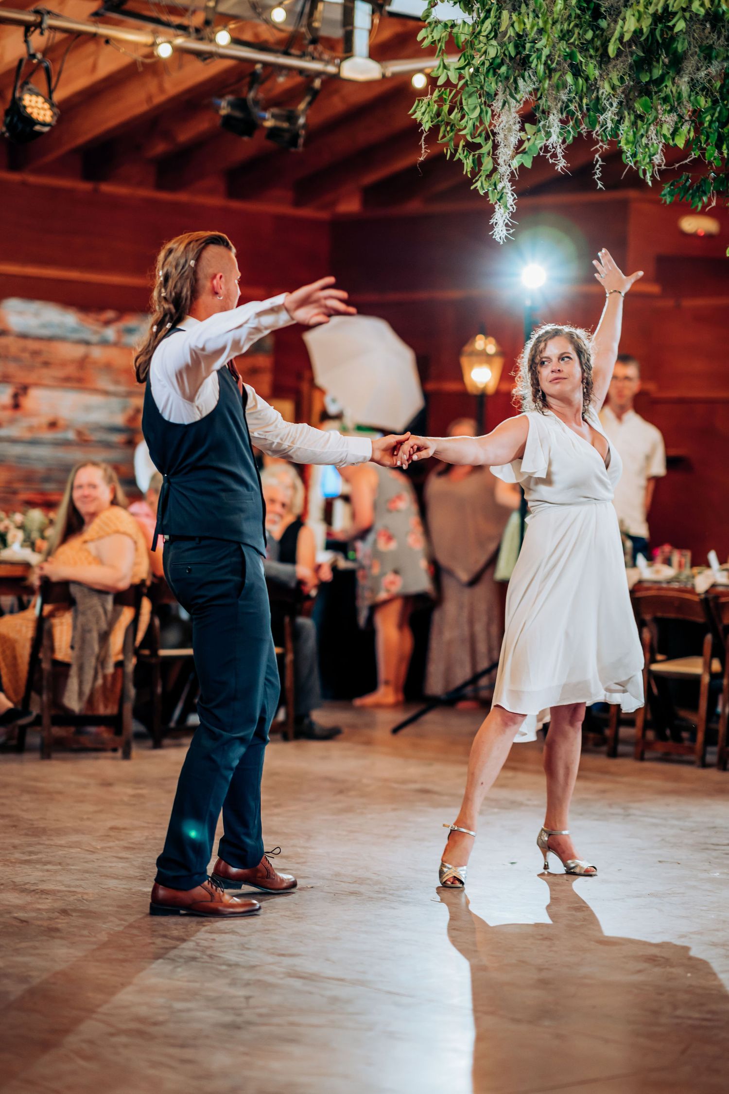 Wedding dancers perform choreographed moves on a dance floor under rustic wooden beams.