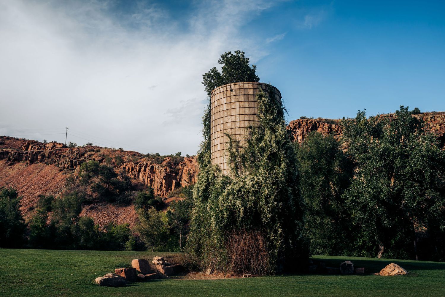 Old stone silo stands against dramatic red cliff landscape under cloudy sky.