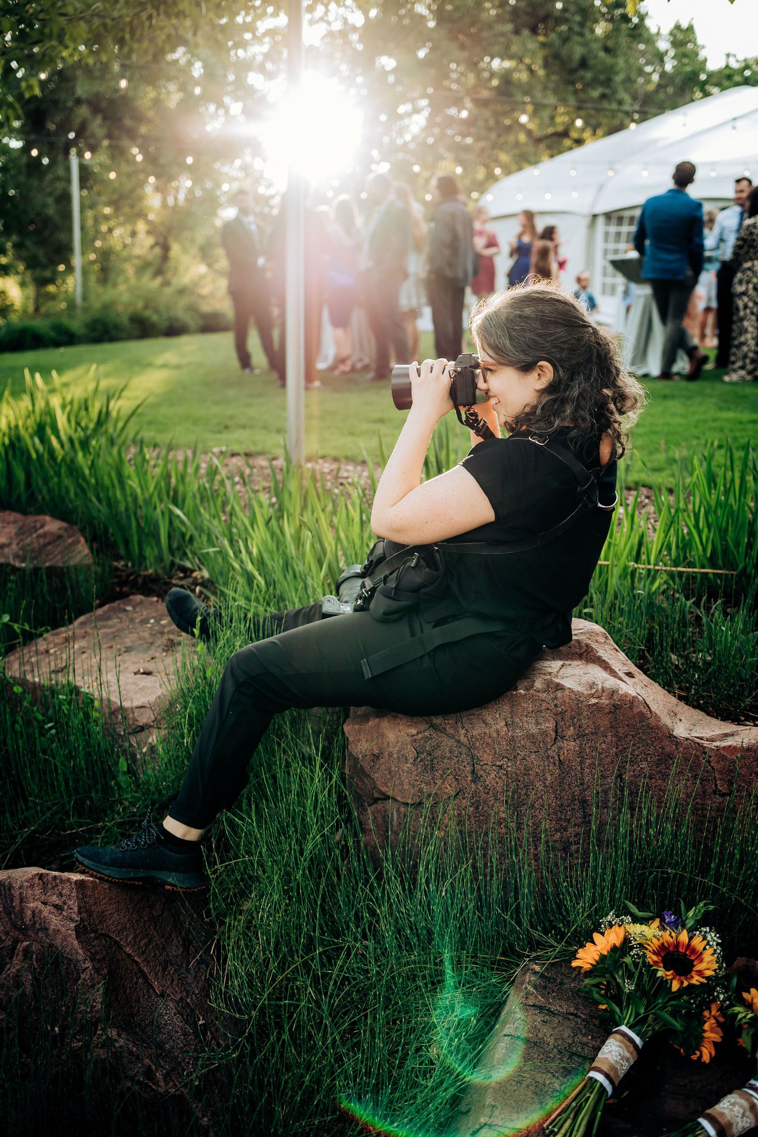 Wedding photographer capturing moments while sitting on rock during outdoor reception.