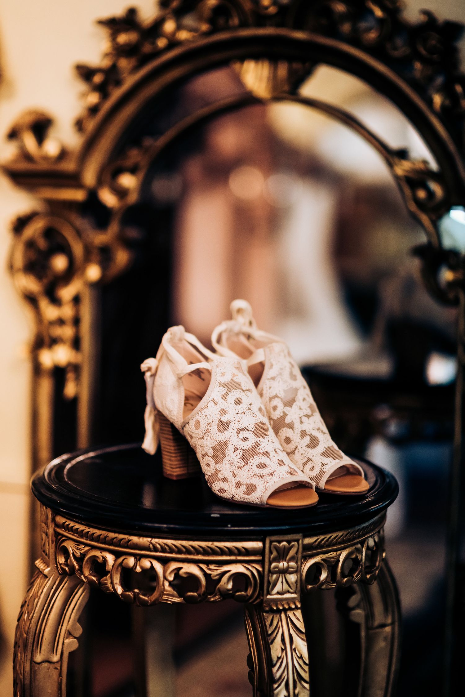 Lace peep-toe wedding shoes displayed on an ornate antique table with mirror reflection.