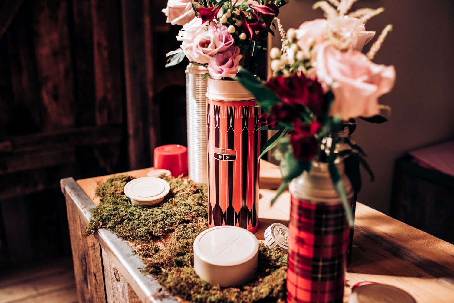 Holiday themed table decor with plaid thermoses, candles and floral arrangements on wooden surface.