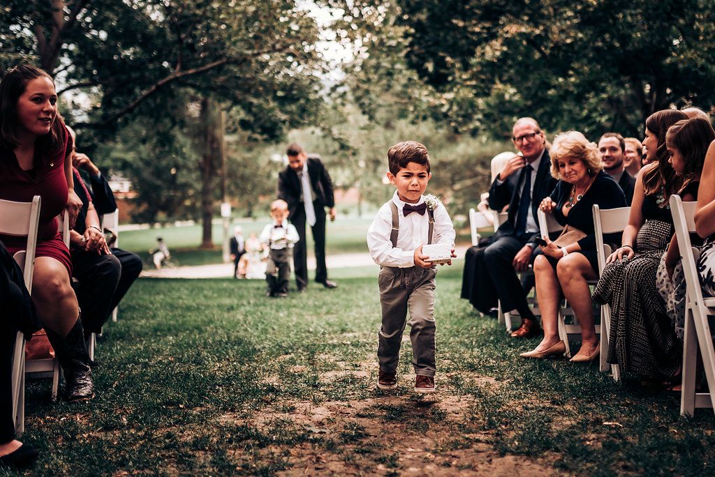 Small child in suspenders and bowtie walking down outdoor ceremony aisle with guests seated on both sides.