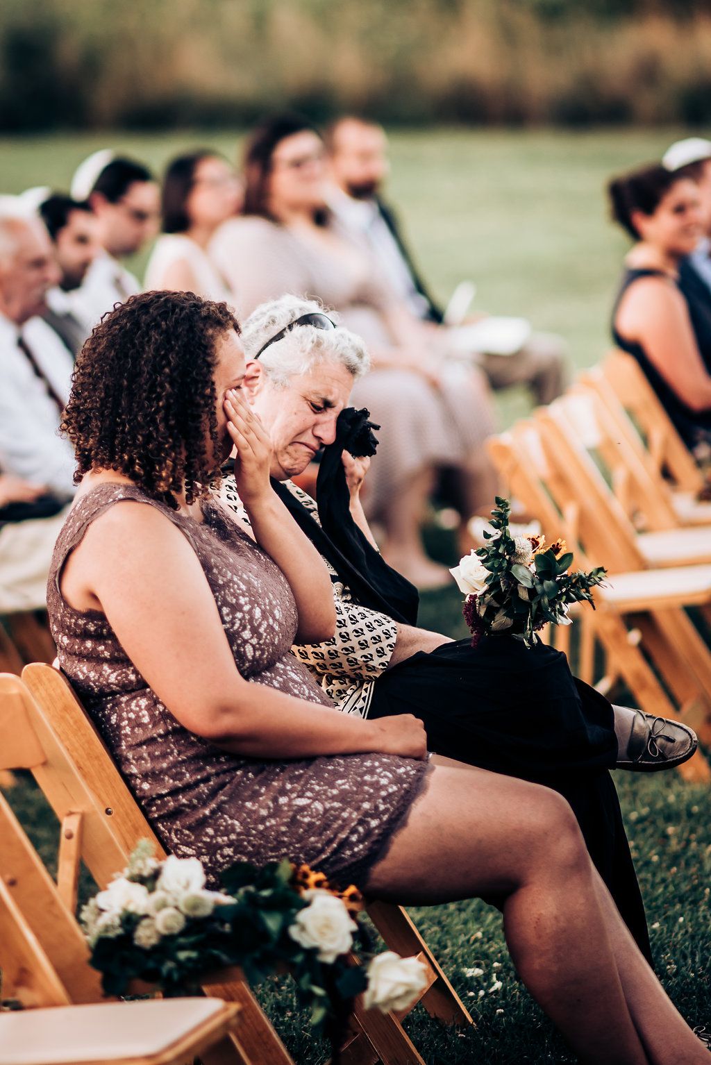 Wedding guests seated on wooden chairs during outdoor ceremony with floral decorations.