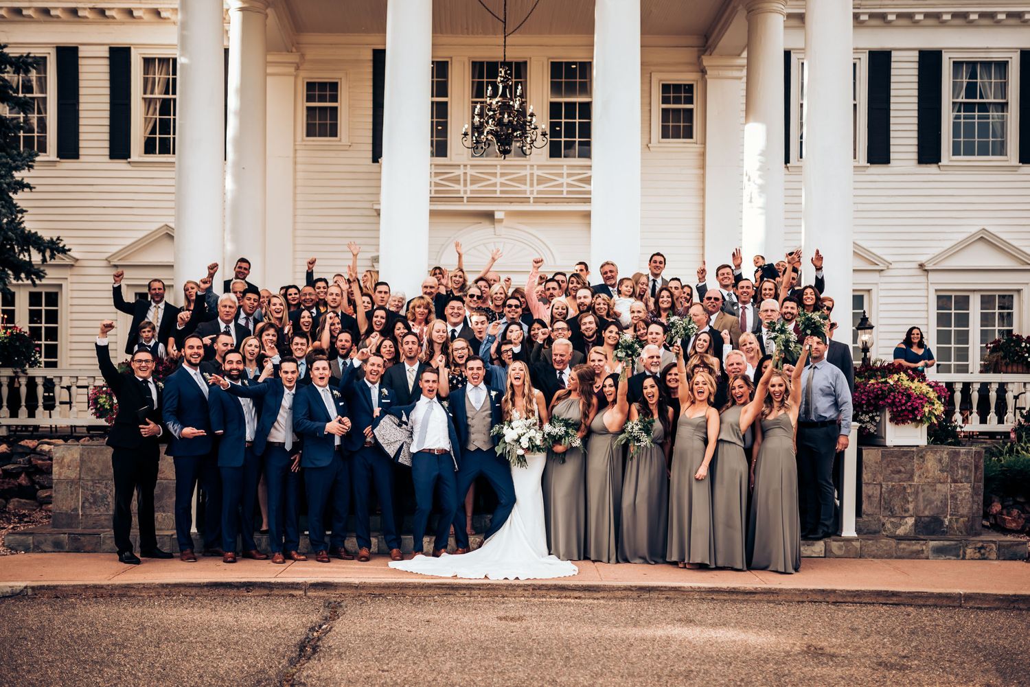 Large wedding party poses for group photo on steps of grand white columned building.