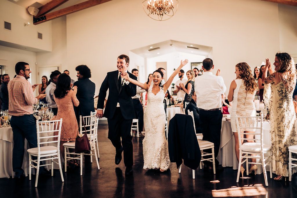 Wedding celebration in a bright reception hall with white chairs and wooden beams.