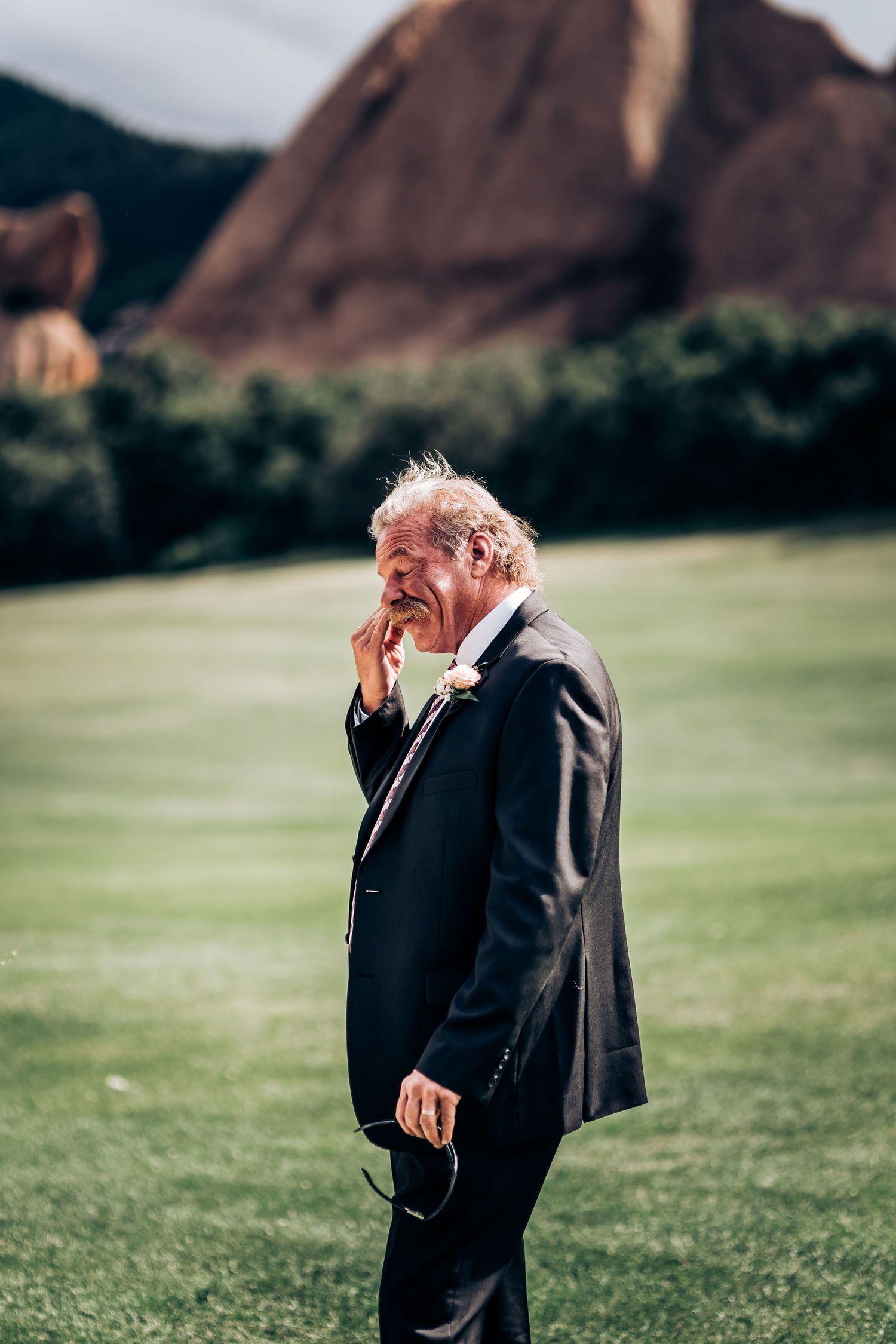 Emotional man in dark suit stands outdoors with red rock formations in background.