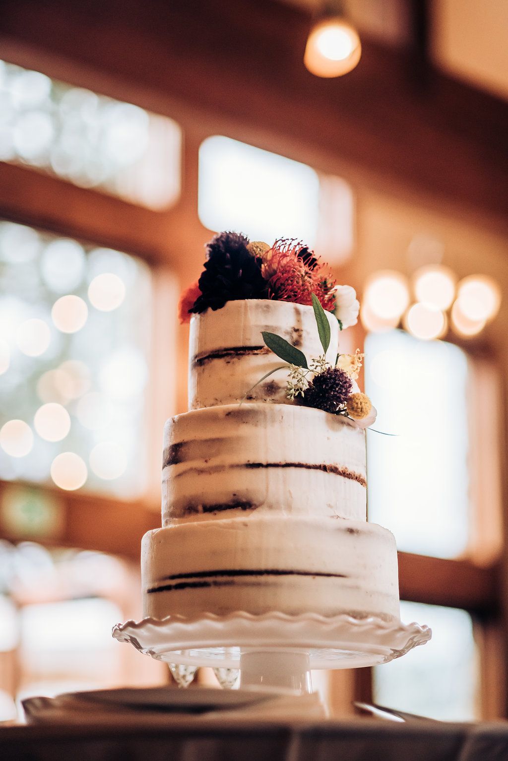 Semi-naked wedding cake with dark floral accents and natural greenery against blurred bokeh background.