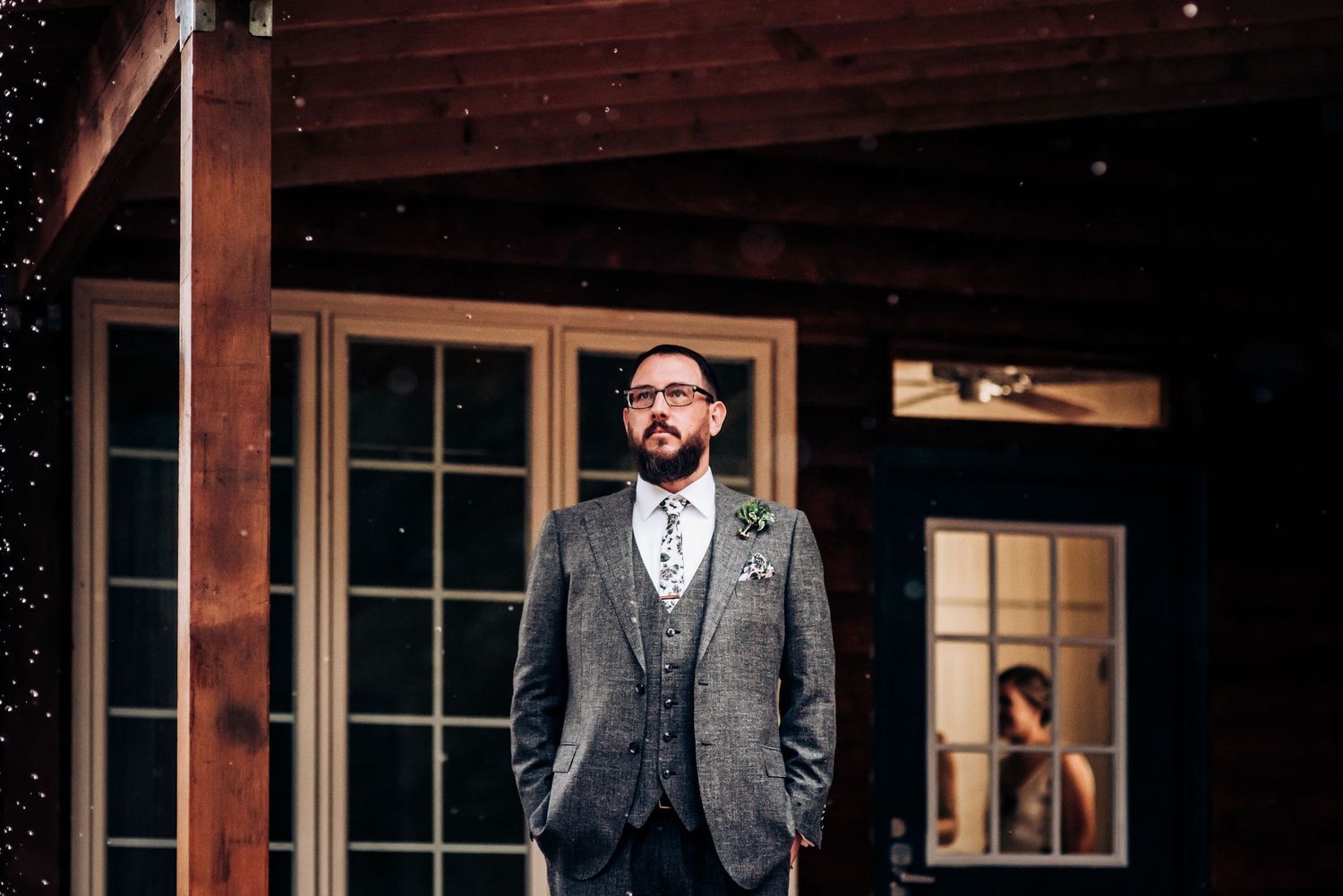 Groom in grey suit stands on porch of rustic cabin during first look photos.