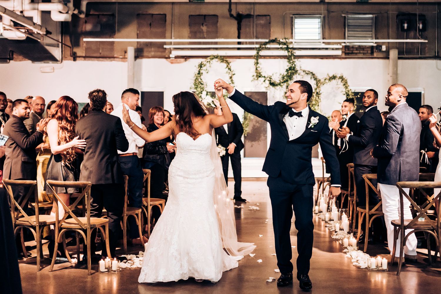 Celebrating couple walks down aisle at indoor wedding ceremony with floral arch backdrop.