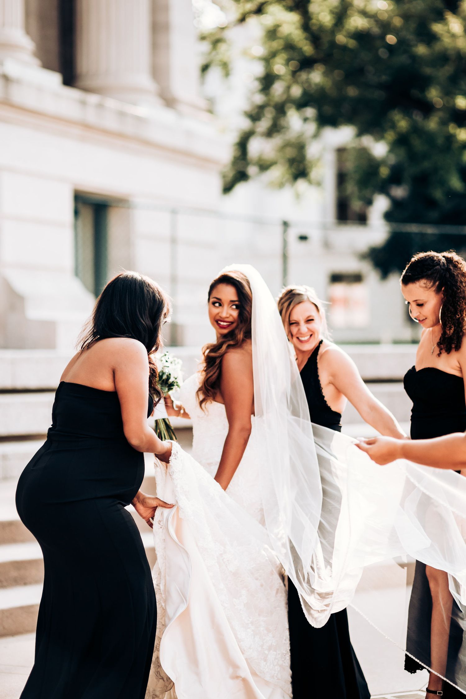 Bridesmaids in black gowns help arrange the bride's veil on outdoor steps.