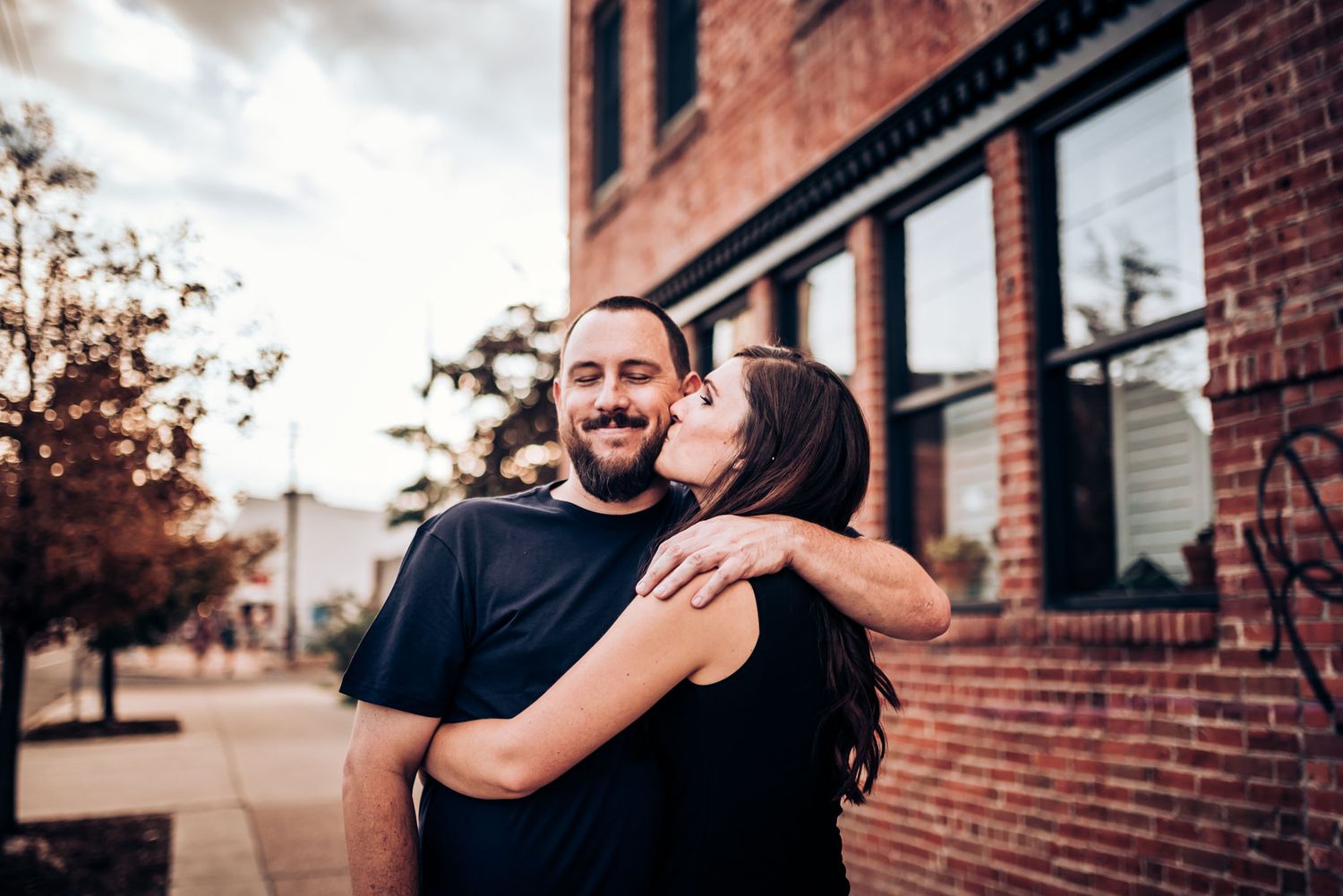 A couple embraces and smiles in front of a brick building during sunset.