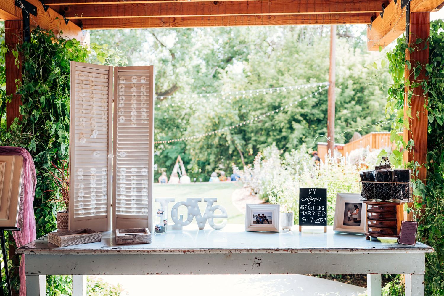 Rustic outdoor wedding display table with shutters and frames under wooden pergola.