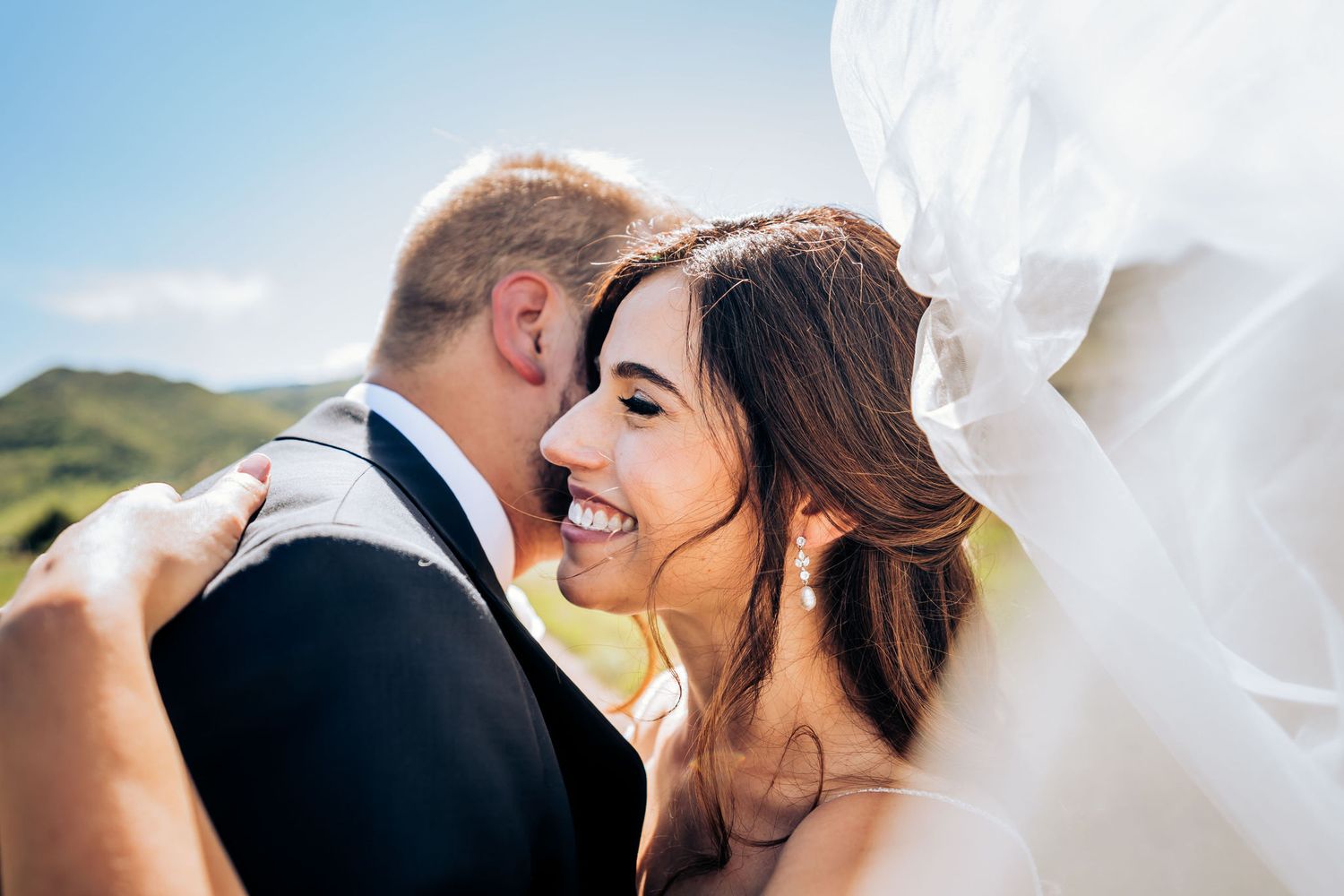 Joyful newlyweds share a candid moment outdoors with flowing white veil in sunlight.