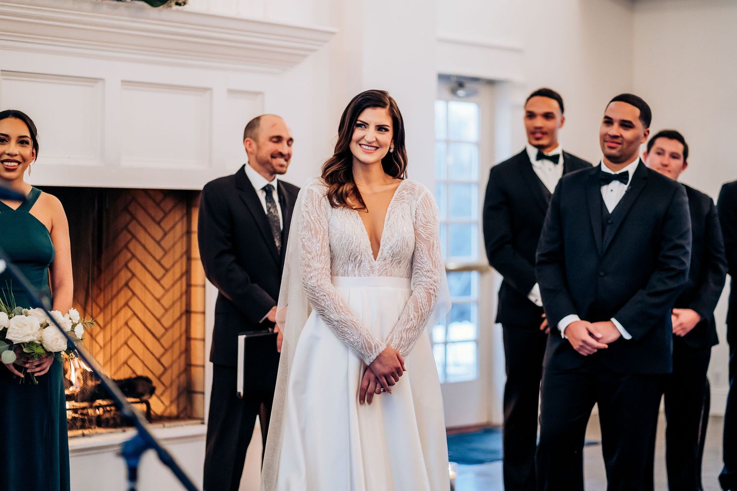 Wedding party stands at front of ceremony space with elegant white decor and fireplace.