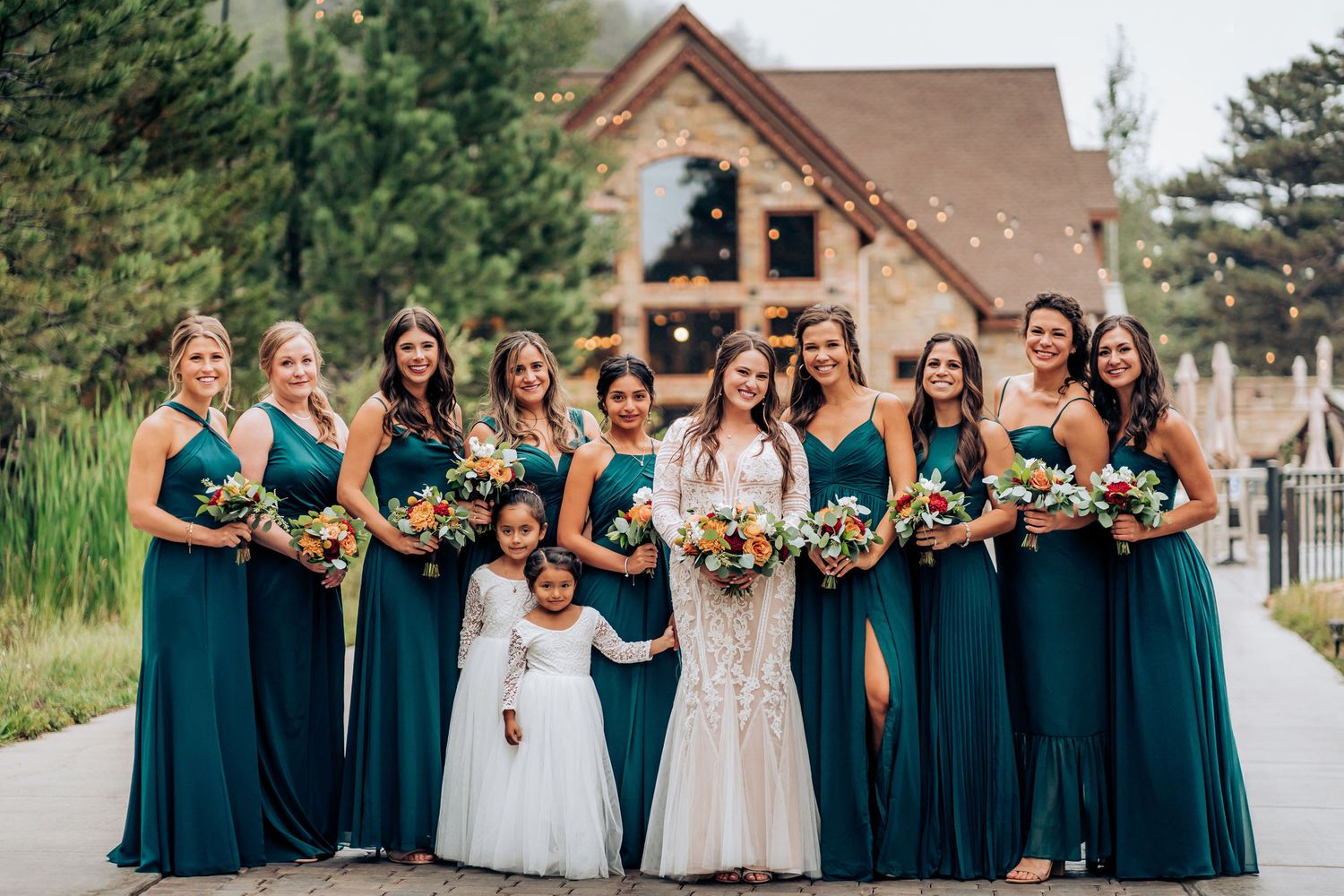 Bridal party in teal dresses poses with flower girl in white dress against rustic lodge backdrop.