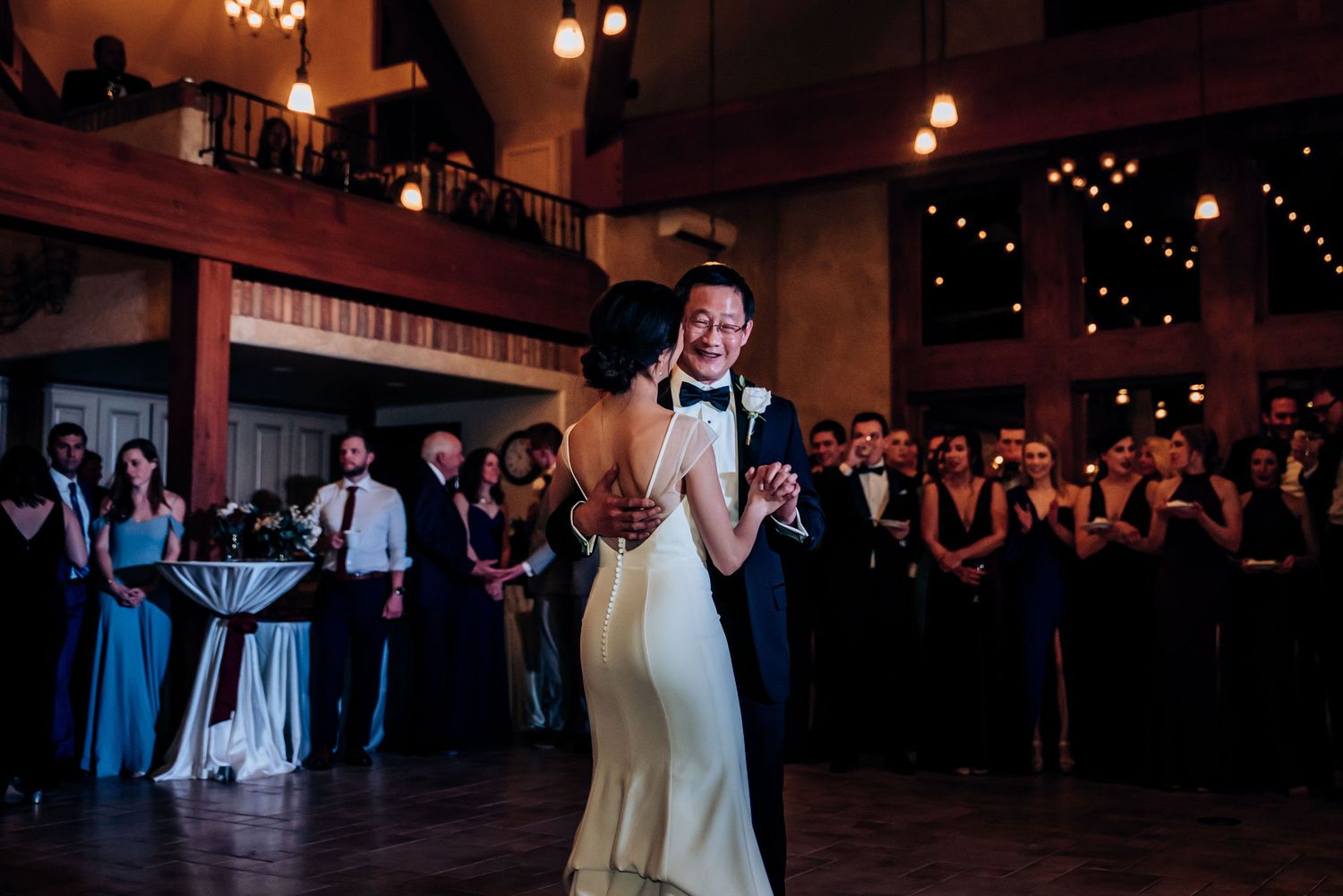 A traditional wedding dance scene in a rustic venue with watching guests and wooden balconies.