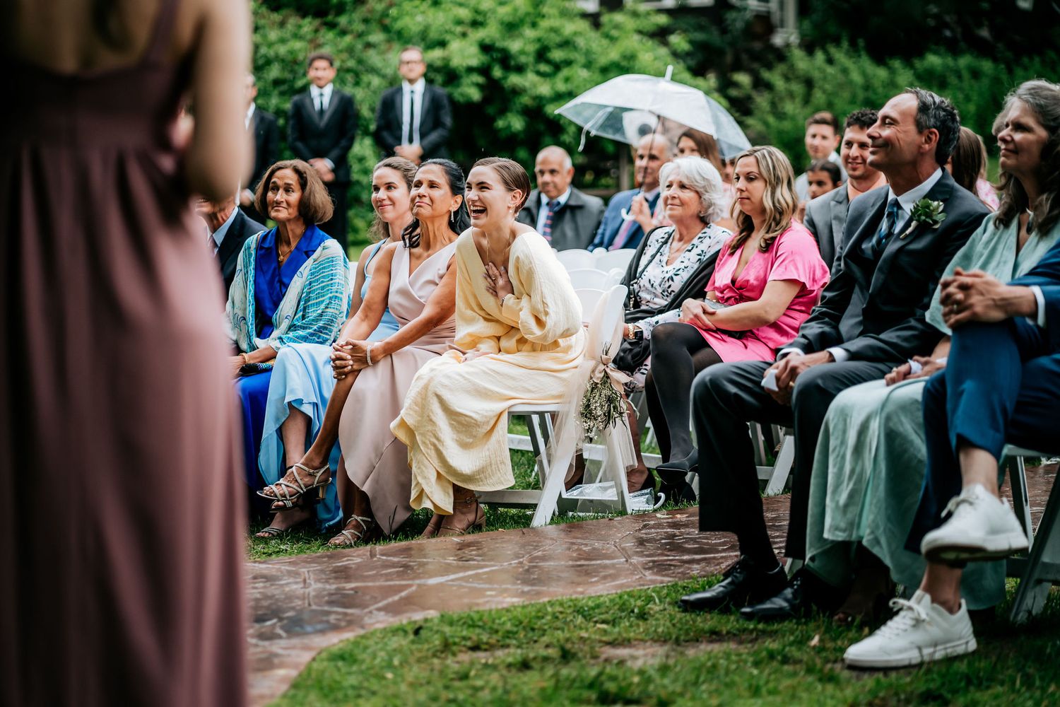 Wedding ceremony guests seated outdoors on white chairs holding umbrellas during light rain.