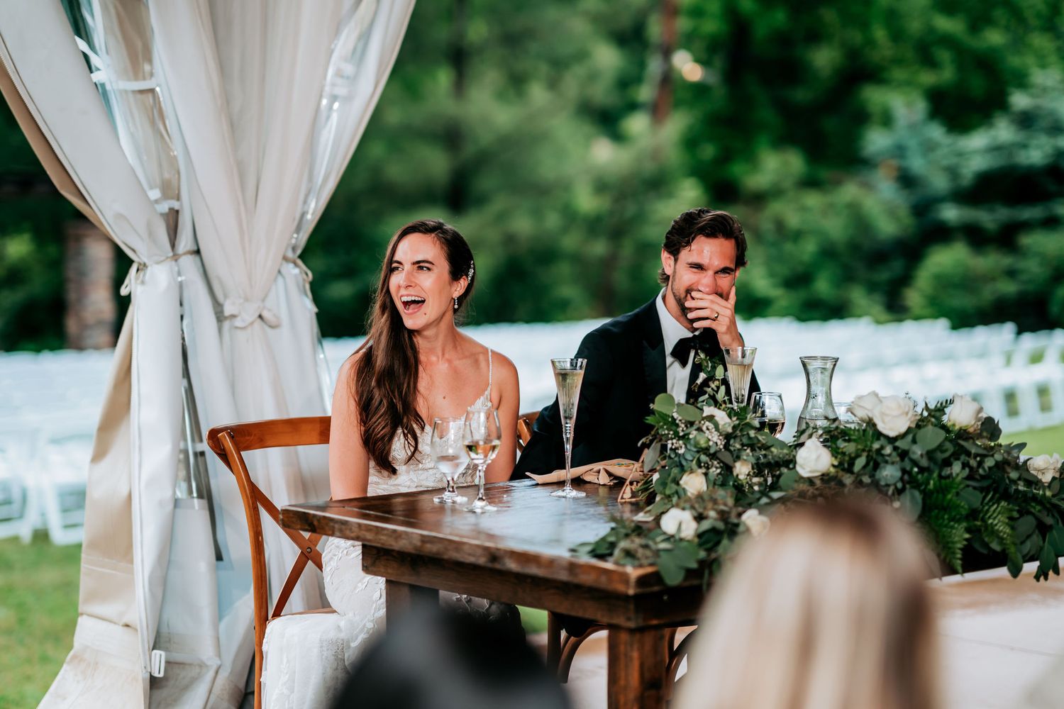 Couple sits at decorated sweetheart table during outdoor wedding reception.