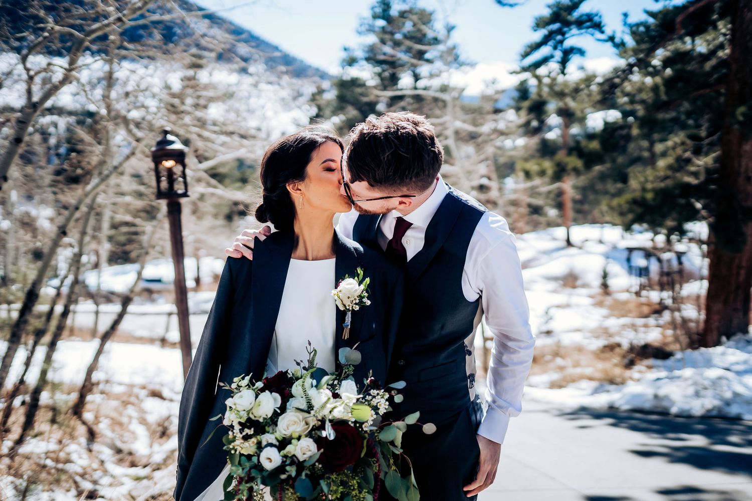 A romantic winter wedding moment captured in a snowy mountain setting with pine trees and a beautiful bouquet.