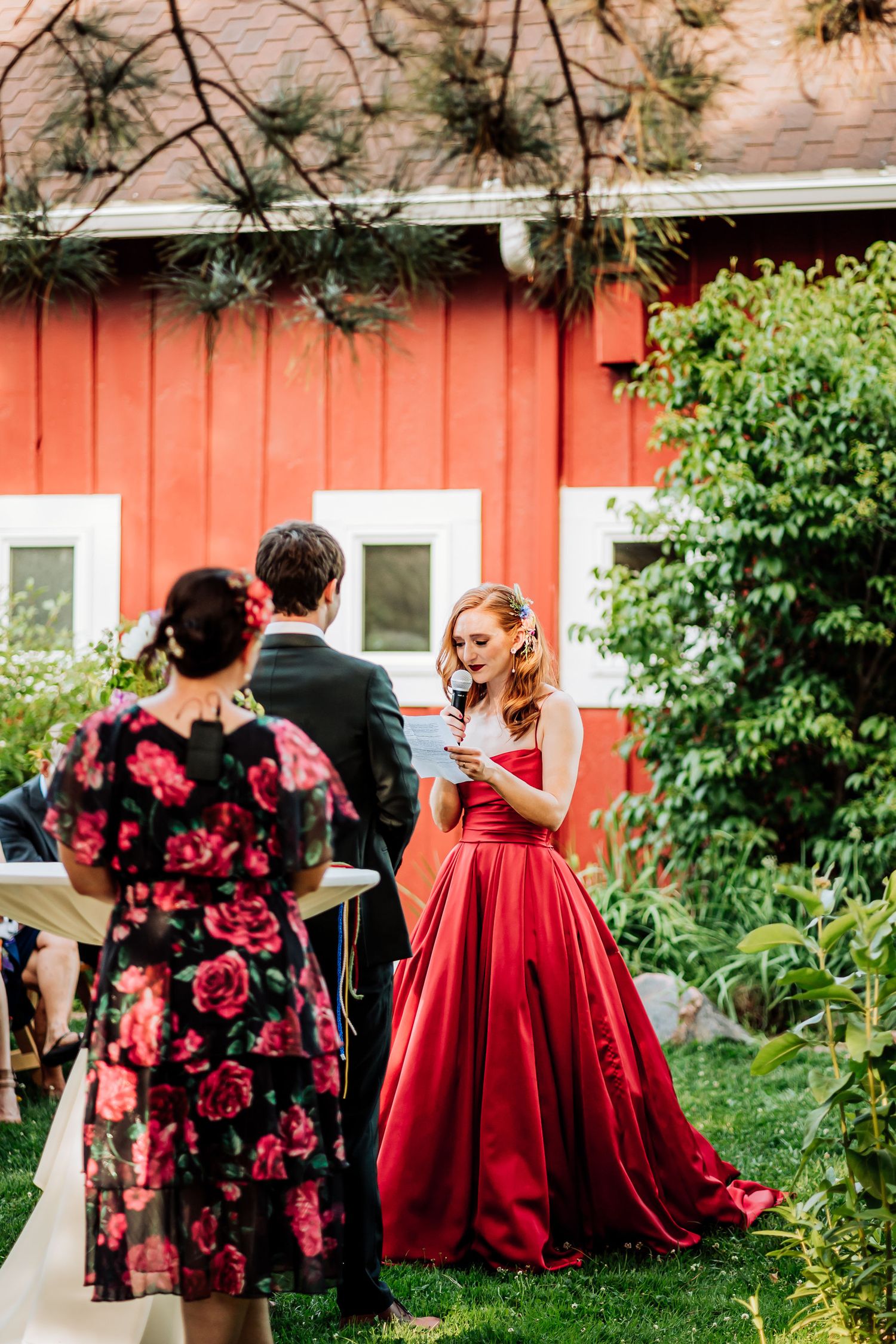 A wedding ceremony takes place outside a rustic red barn with guests in floral dresses.