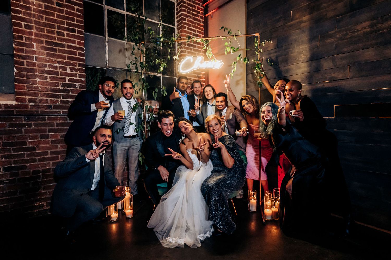 Large wedding party poses together for group photo in front of brick wall with decorative lighting.