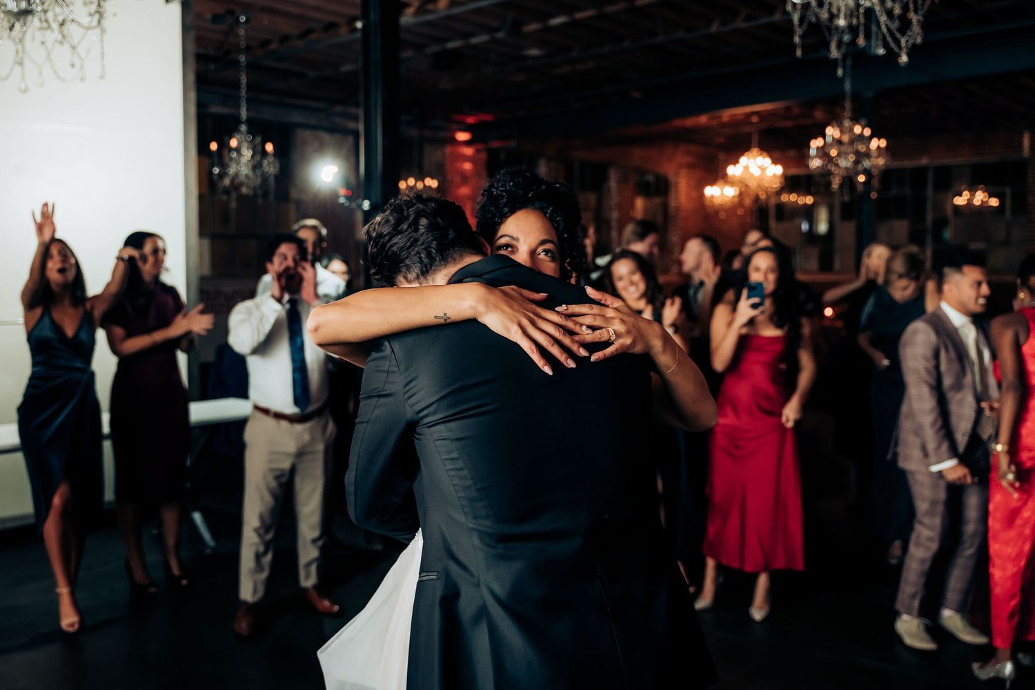 Wedding guests dance and embrace on a dimly lit dance floor with chandeliers overhead and red accent lighting.