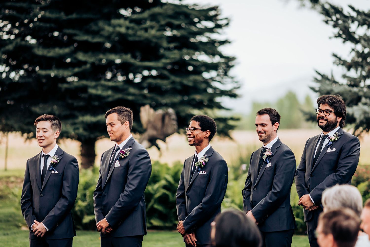 Groomsmen in navy suits stand in line during outdoor wedding ceremony with evergreen trees.