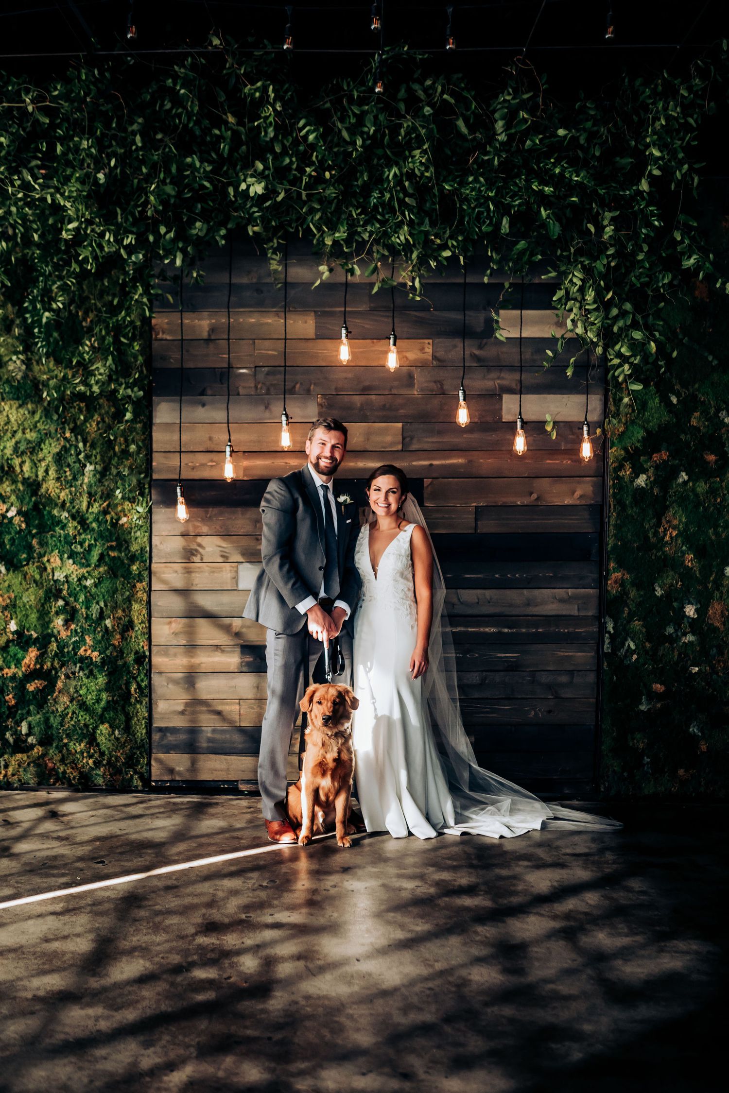 Wedding couple poses with their dog against a rustic wooden wall with hanging lights and greenery.