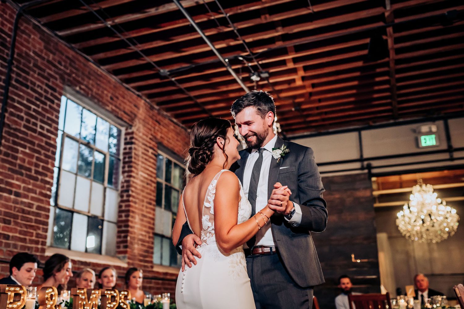 A romantic first dance moment in an industrial brick venue with exposed wooden beams and chandeliers.