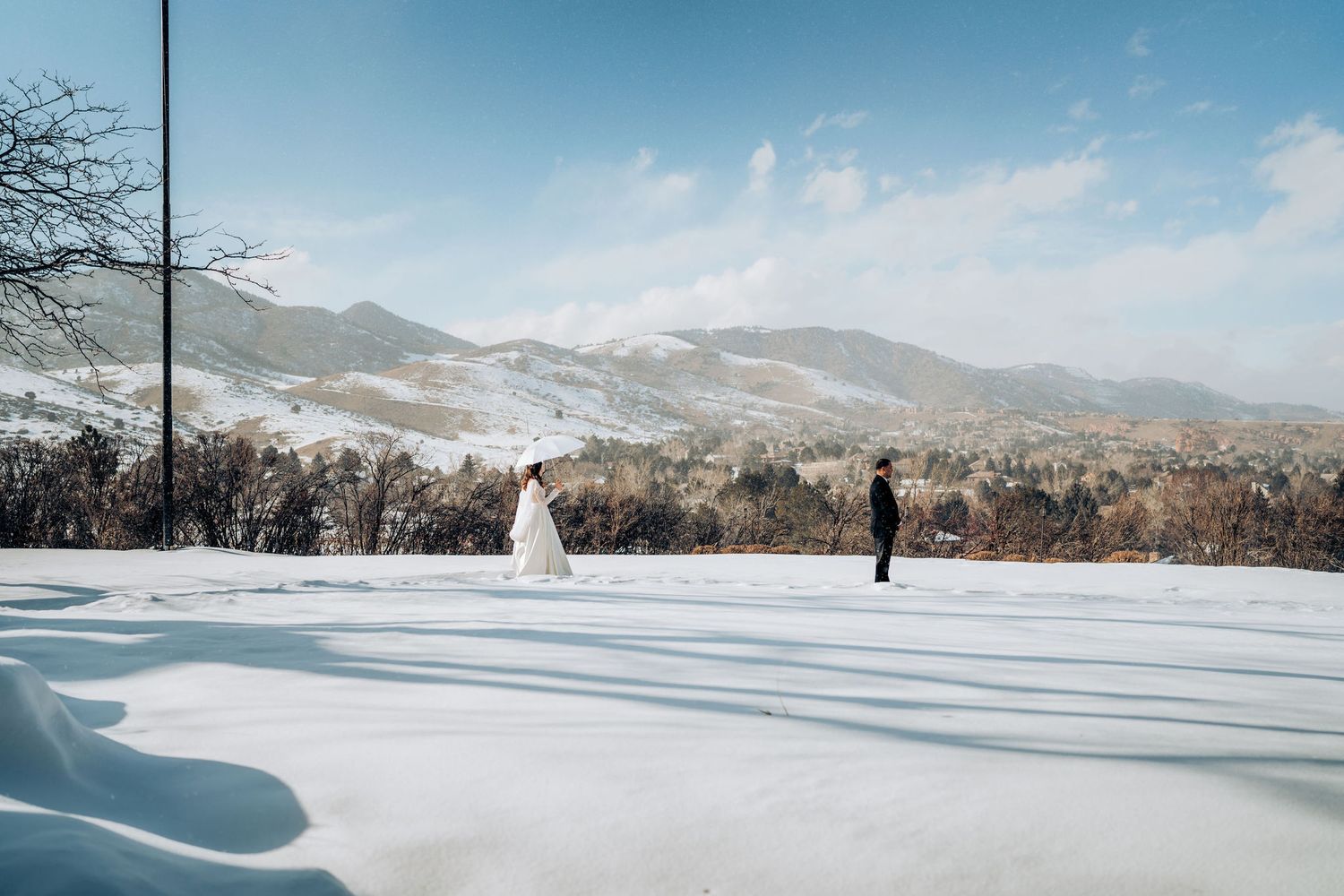 Two figures stand apart in the snow with mountains and bare trees in the background.