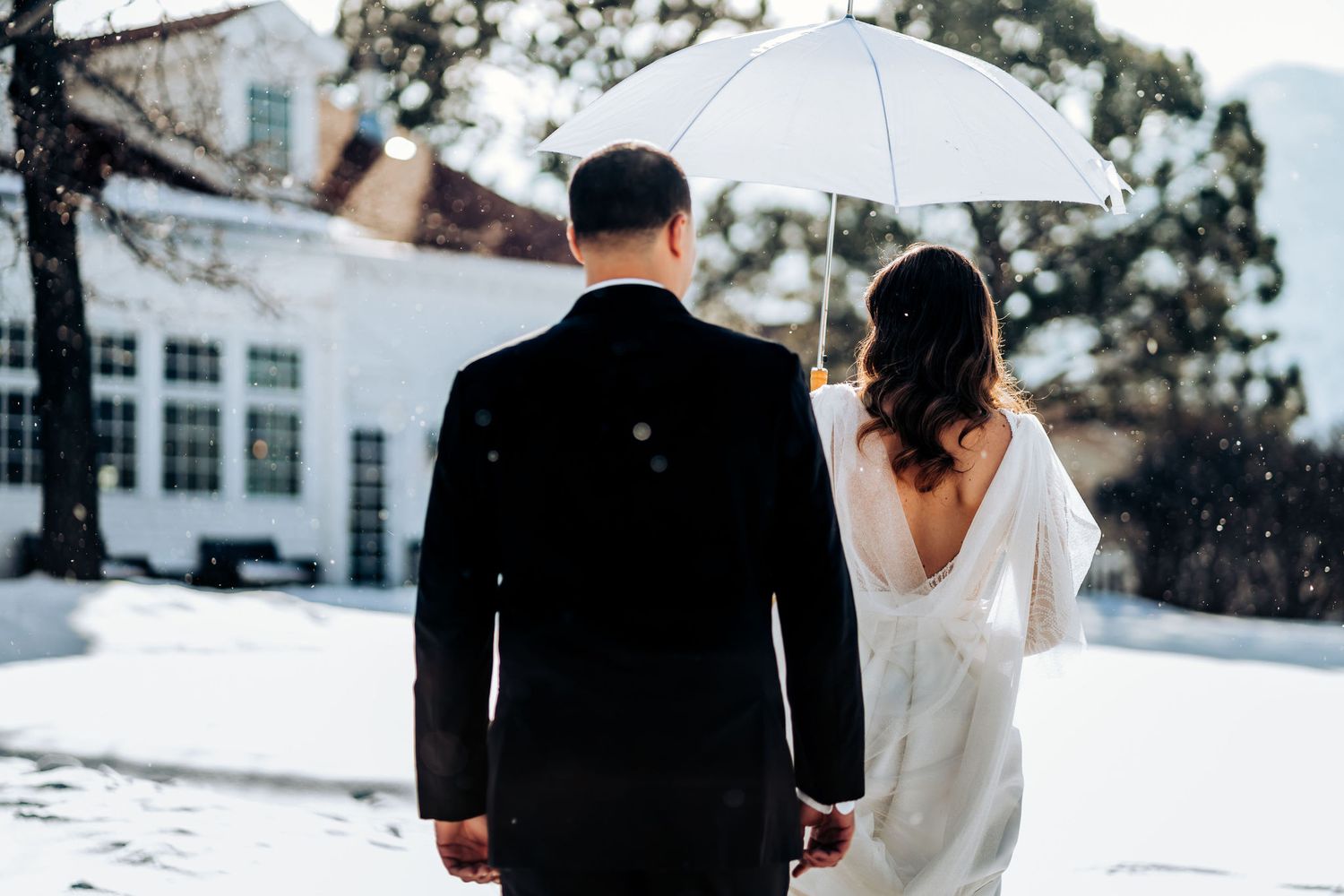Back view of newlyweds holding a white umbrella while standing in snow.
