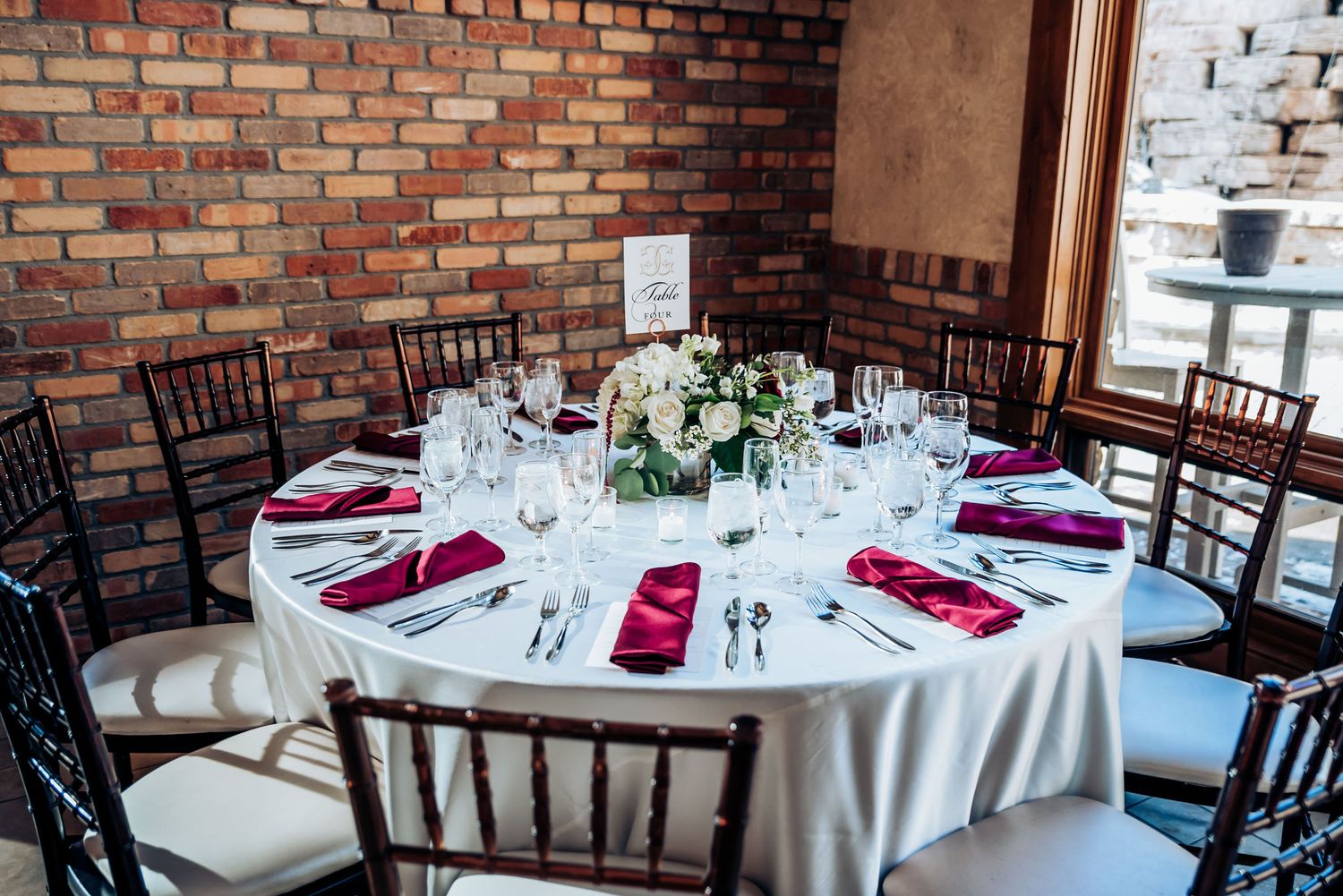 Formal wedding reception table set with burgundy accents against rustic brick wall.