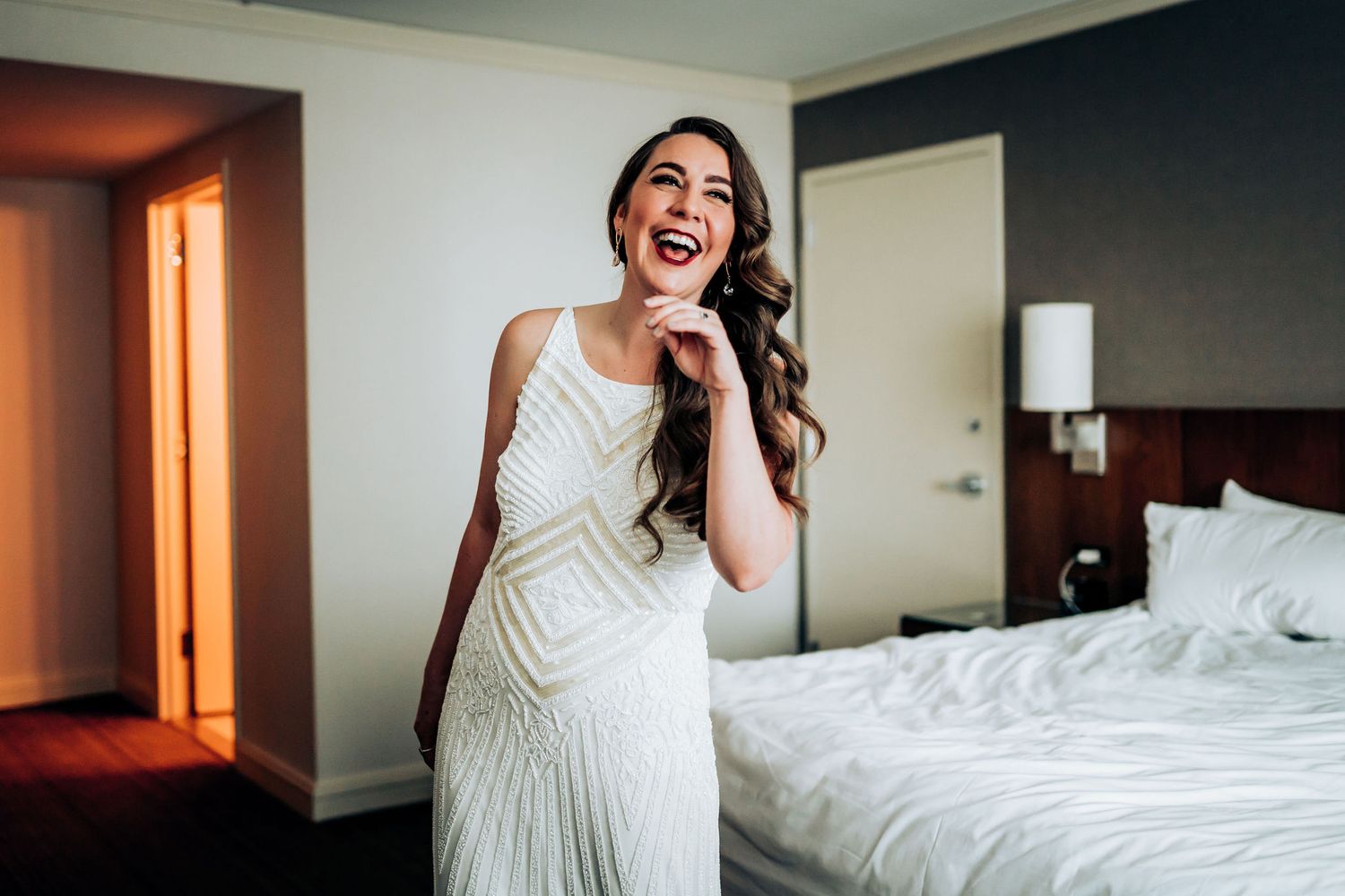 Person in white beaded wedding dress laughing joyfully in a hotel room.
