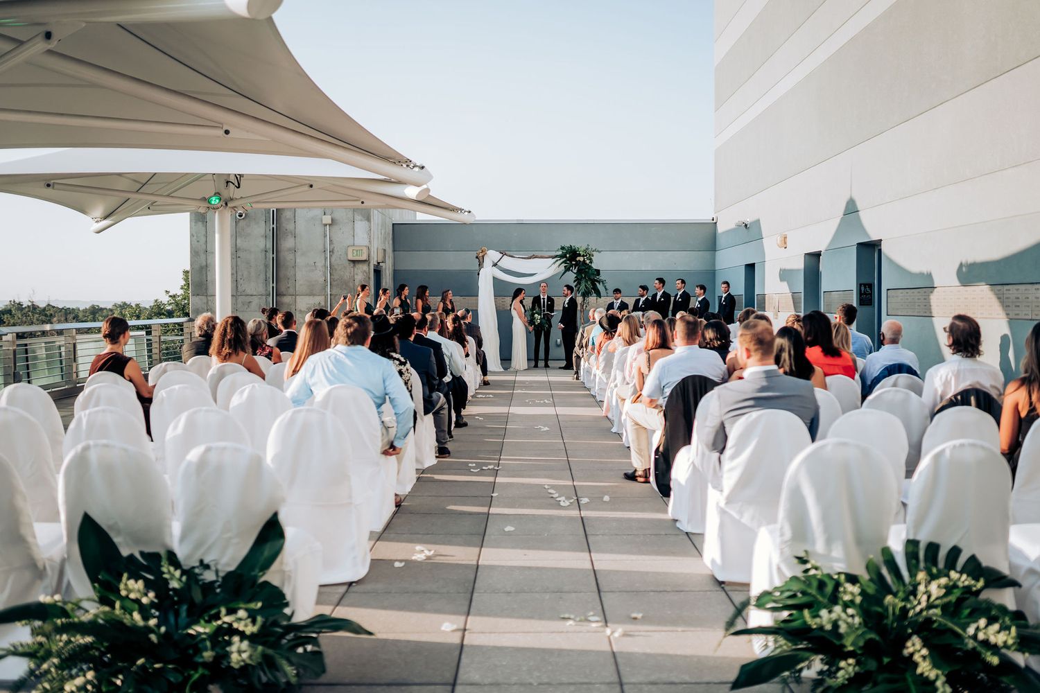 Modern rooftop wedding ceremony with white chairs and floral arch overlooking city views.