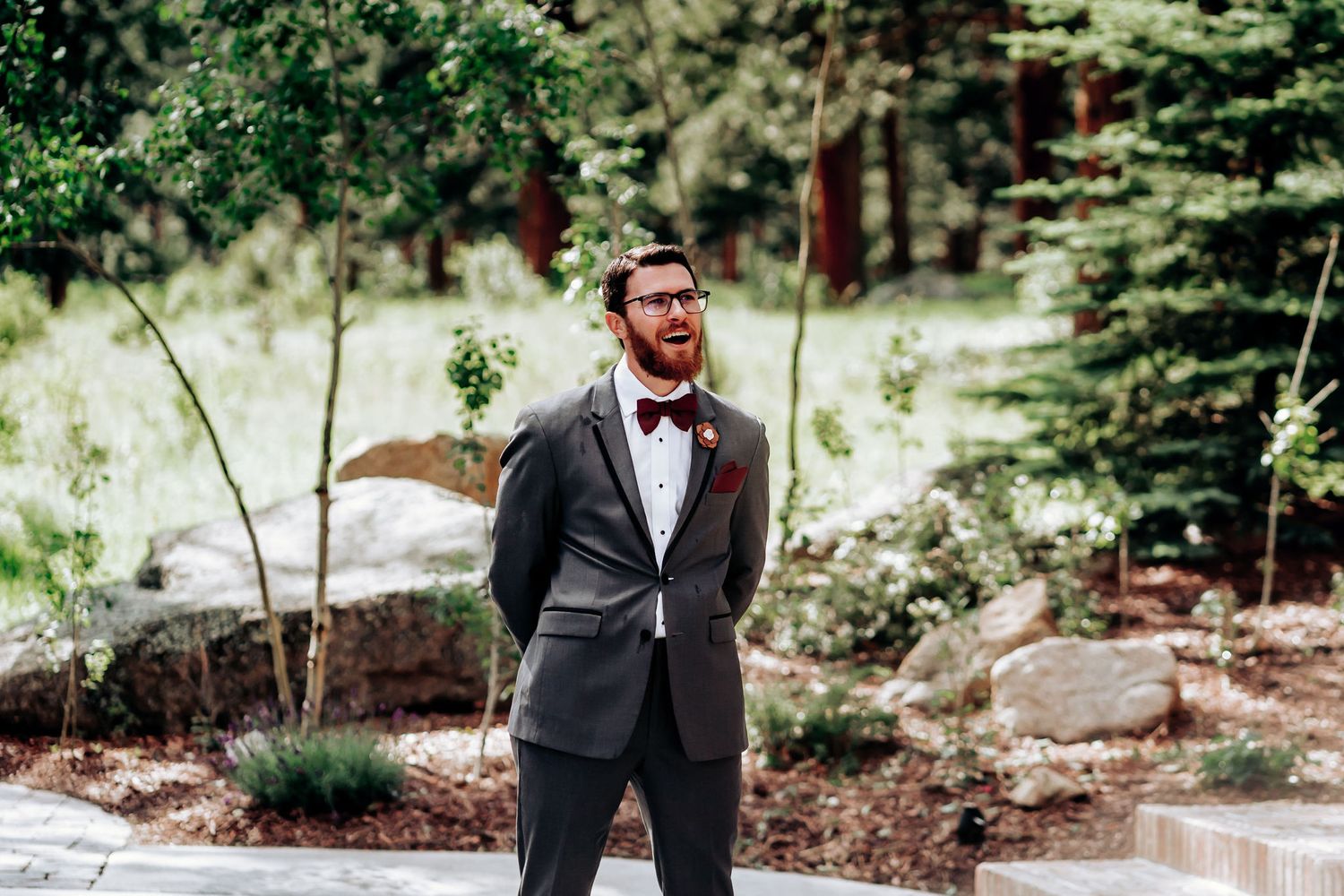 A formal wedding portrait in a natural outdoor setting with evergreen trees and stone landscaping.