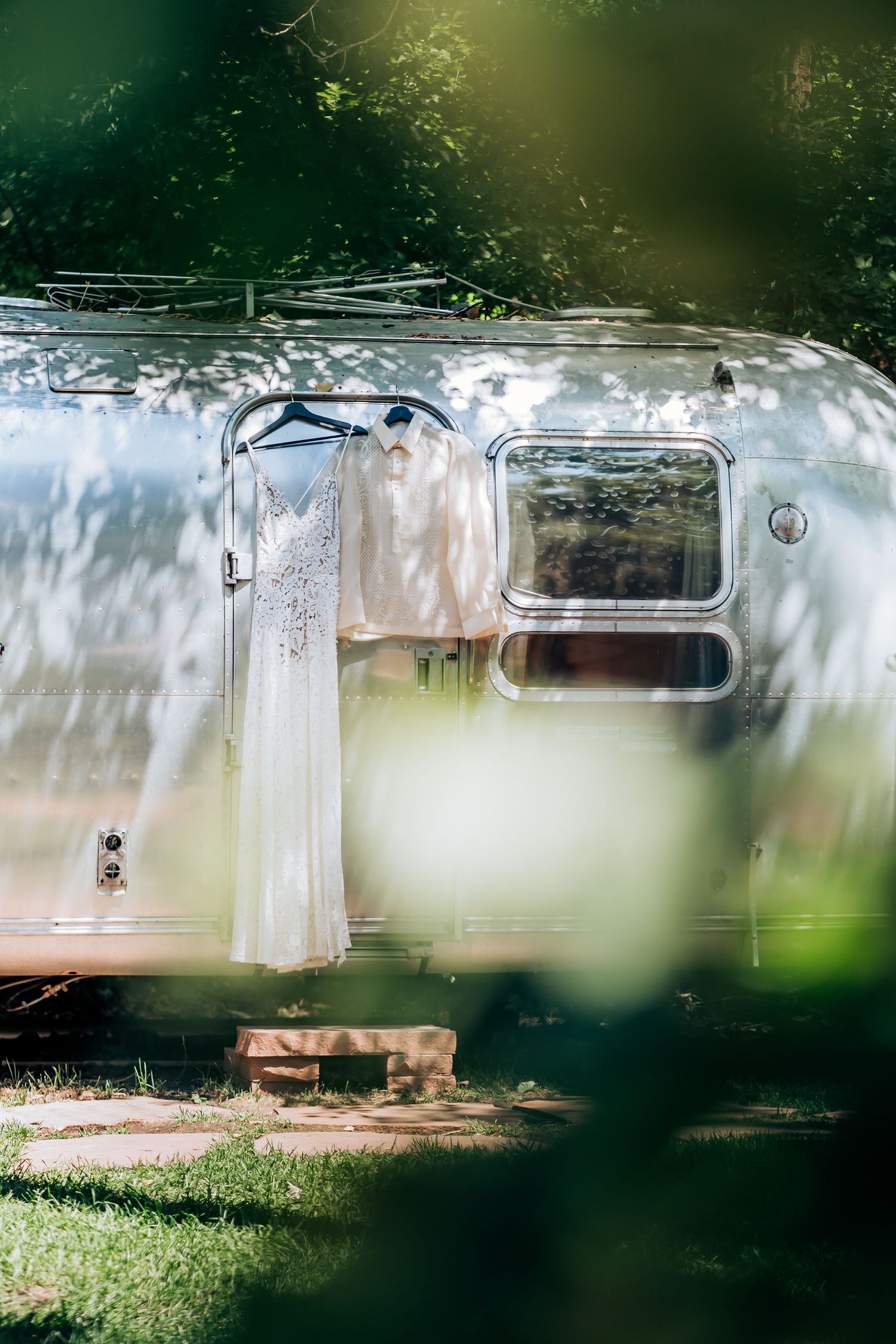 A vintage Airstream trailer with a white wedding dress hanging outside in dappled sunlight.