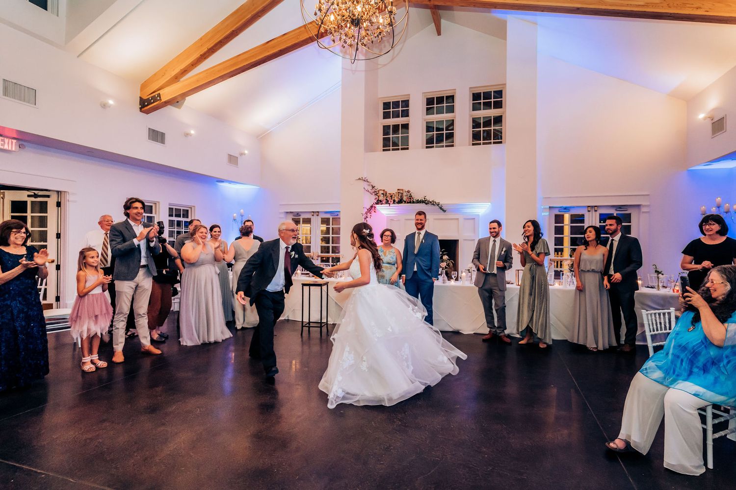Wedding celebration with guests gathered around dance floor in elegant venue with chandeliers.