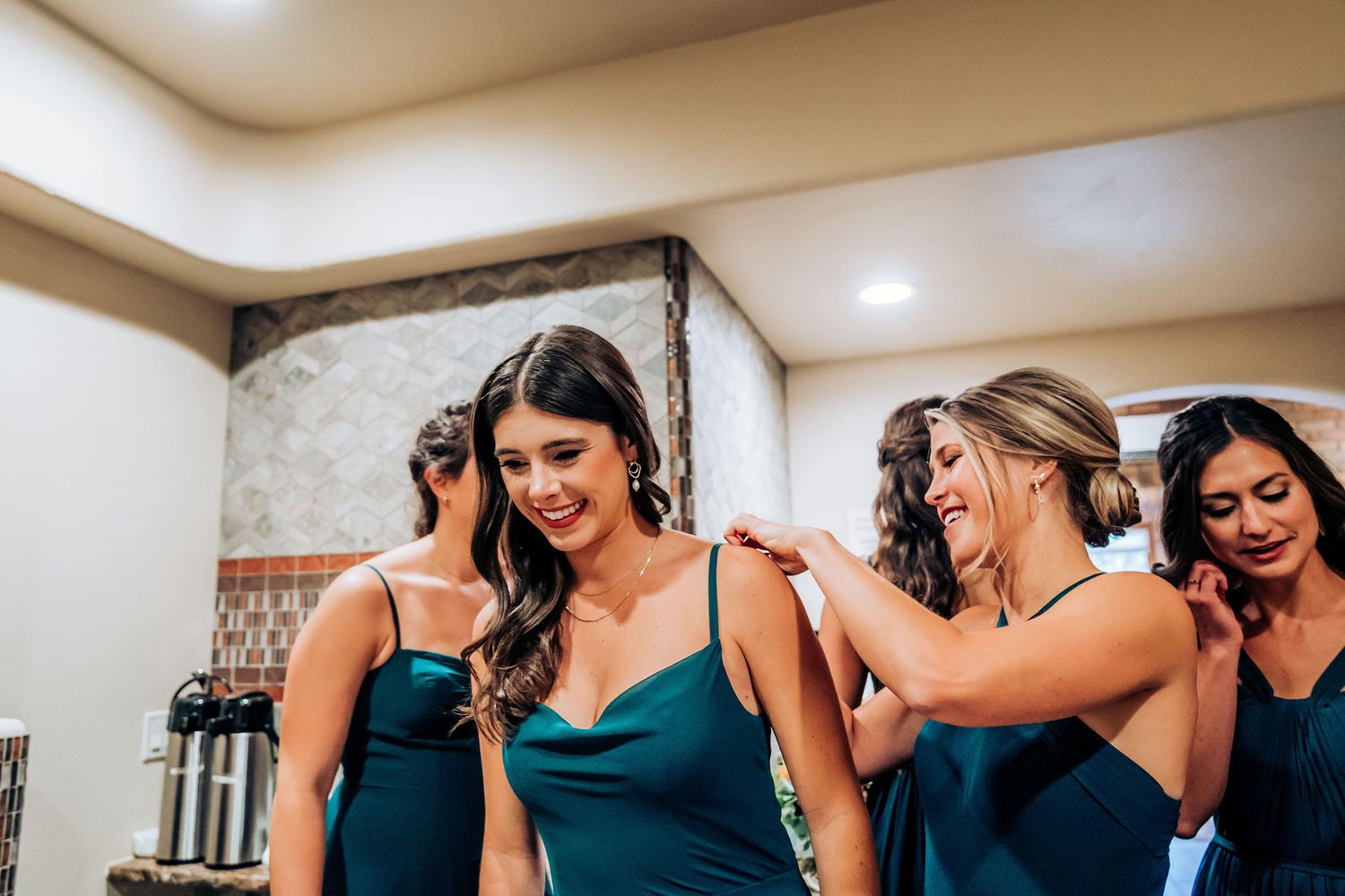 Bridesmaids in teal dresses helping each other get ready in a dressing room.