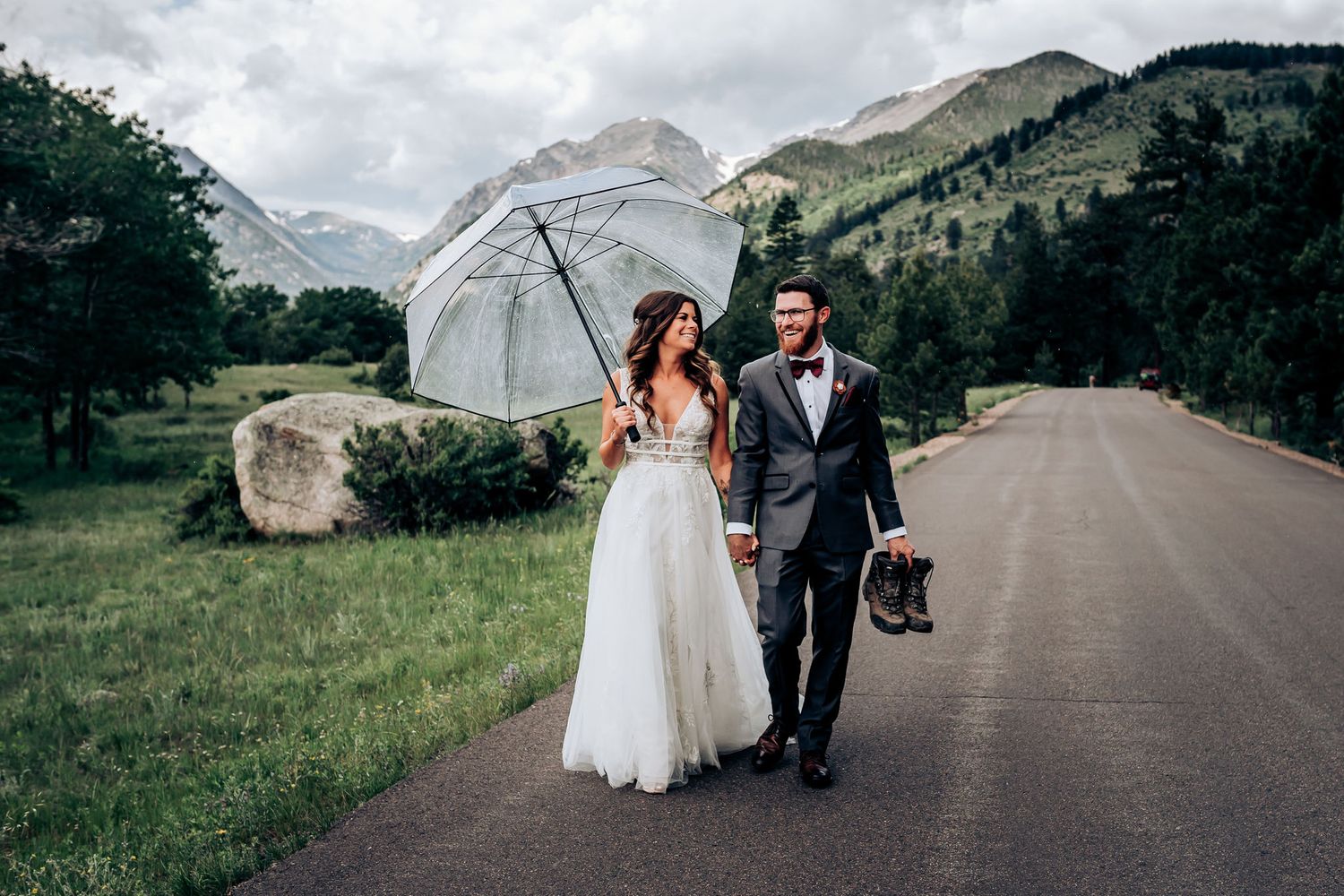 Wedding couple walks along mountain road with white umbrella against scenic backdrop.