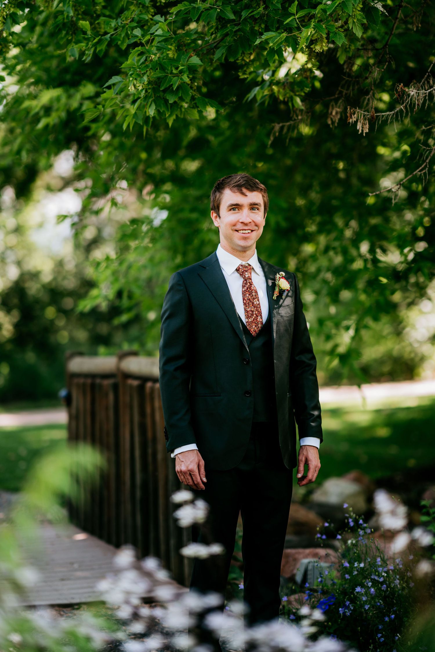 Groom in classic black suit stands on garden bridge surrounded by greenery.