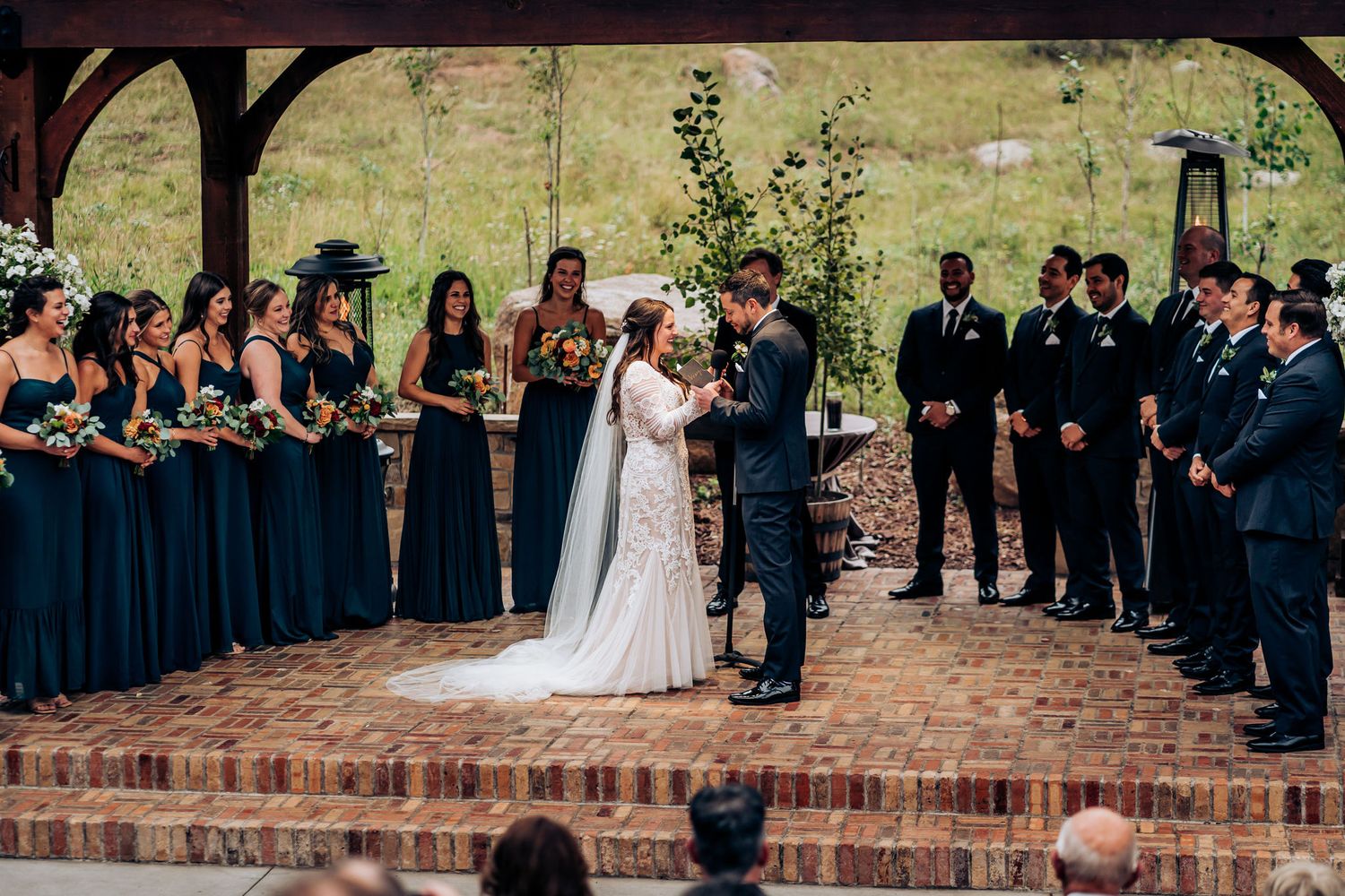 Wedding ceremony under a rustic wooden pergola with wedding party in navy and black attire.