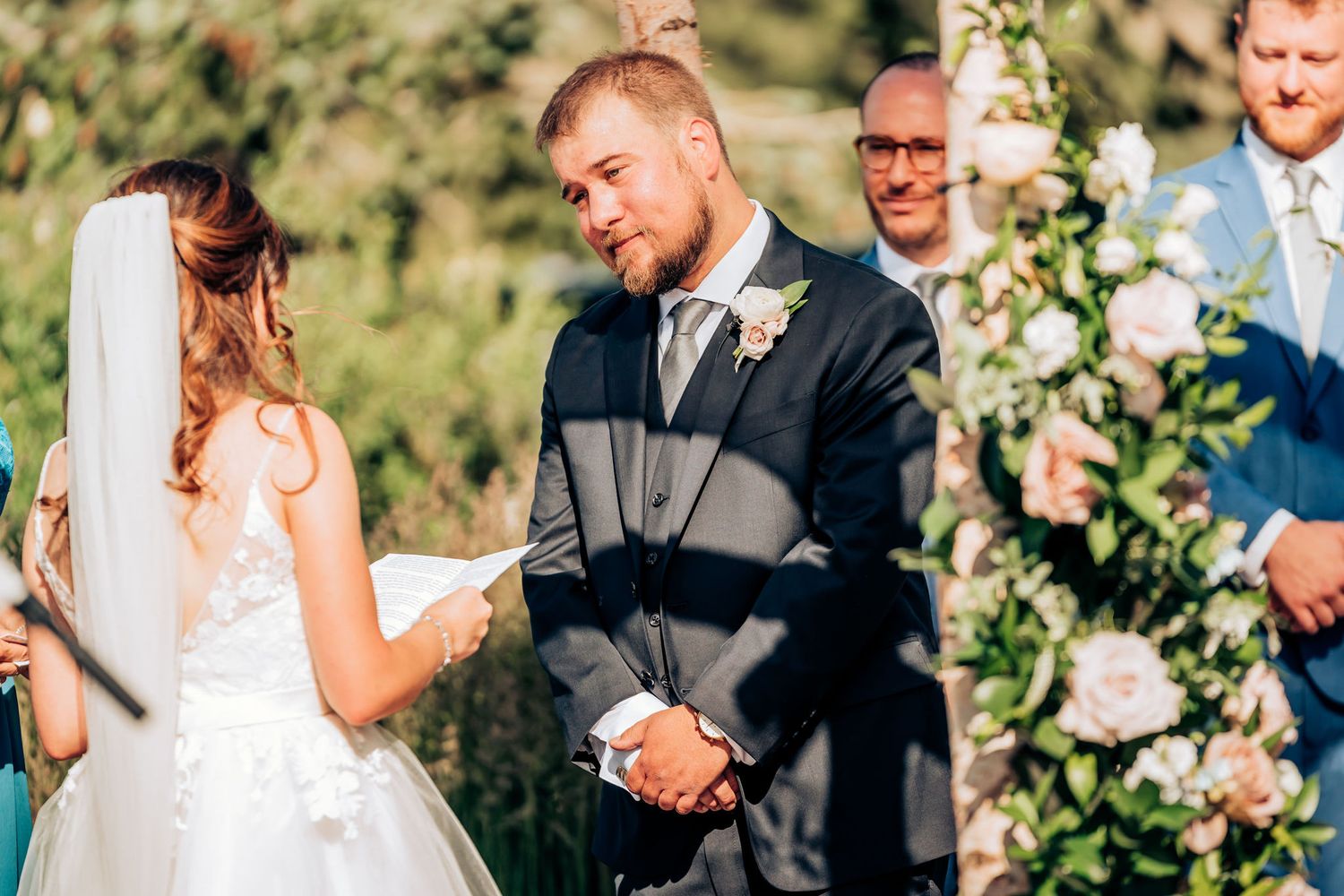 Couple exchanging vows under wooden arch decorated with white flowers during outdoor ceremony.