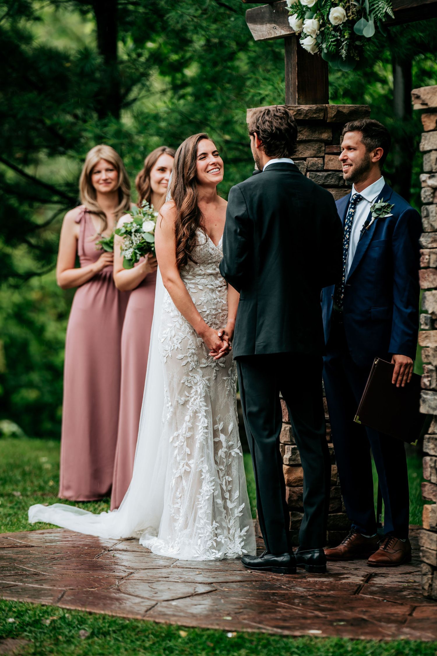 Wedding ceremony at stone arch with bridesmaids in pink dresses and officiant performing service.