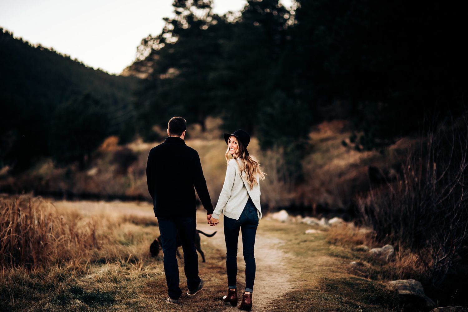 A couple walks hand in hand on a mountain trail at sunset with golden light filtering through trees.