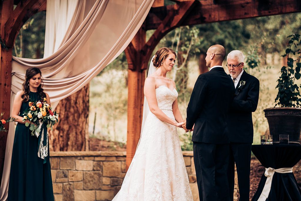 Emotional wedding ceremony taking place under wooden pergola with flowing fabric drapes and stone base.