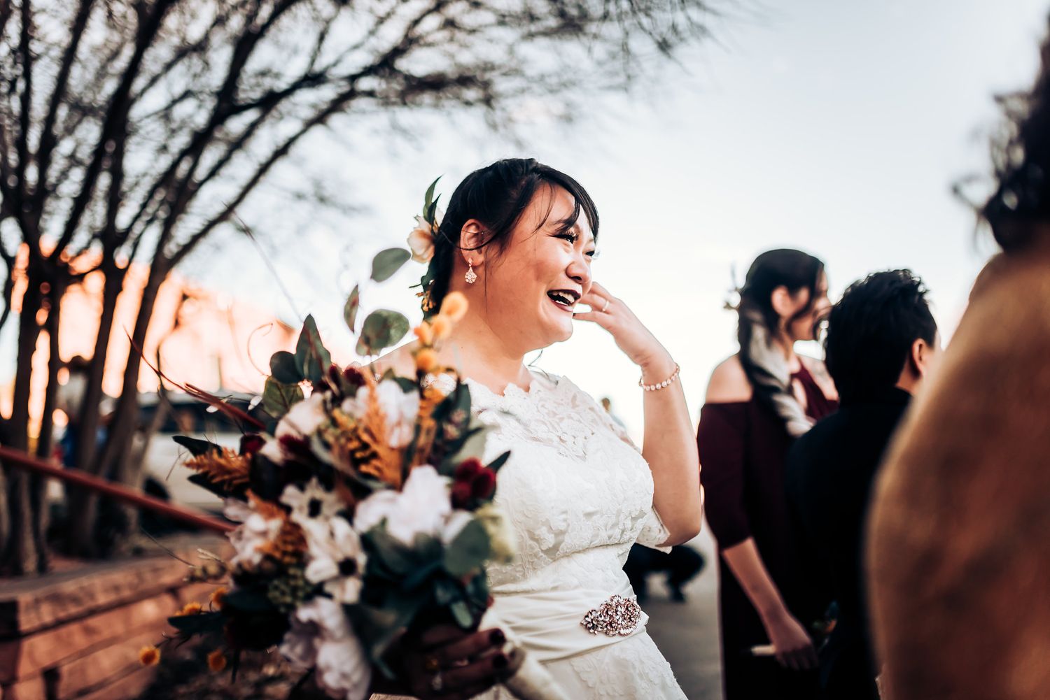 Outdoor wedding celebration with emotional candid moment in white lace dress against sunset sky.