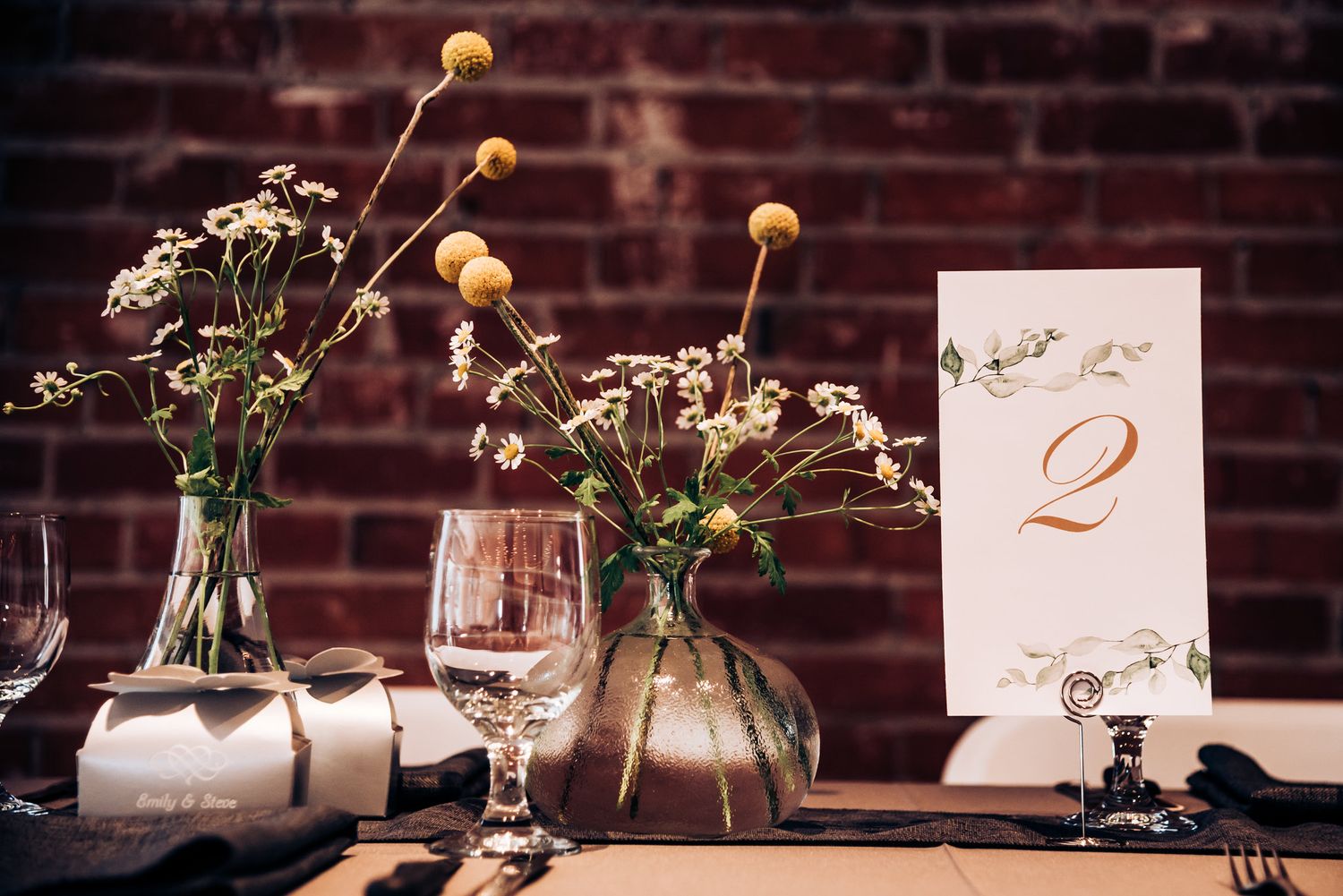 Wedding table decor with yellow flowers, wine glasses and table number against brick wall.