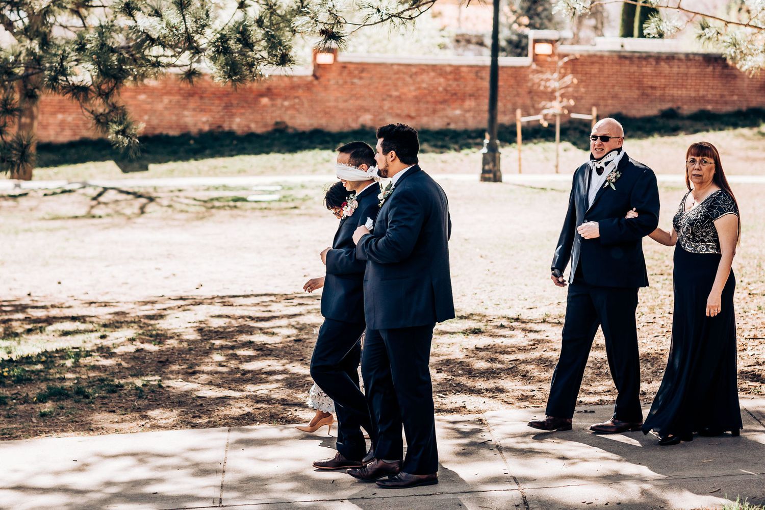 Wedding party members in formal attire gather outdoors near a brick wall on a sunny day.